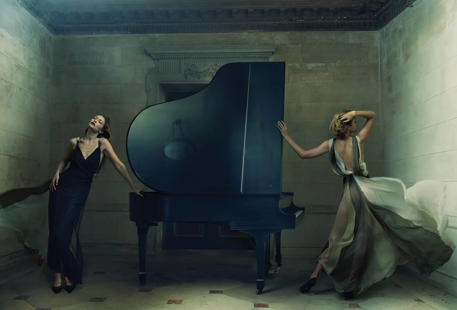 Two women in flowing gowns pose with a grand piano in a classic interior featuring stone walls, arches, and a tiled floor, under soft lighting.
