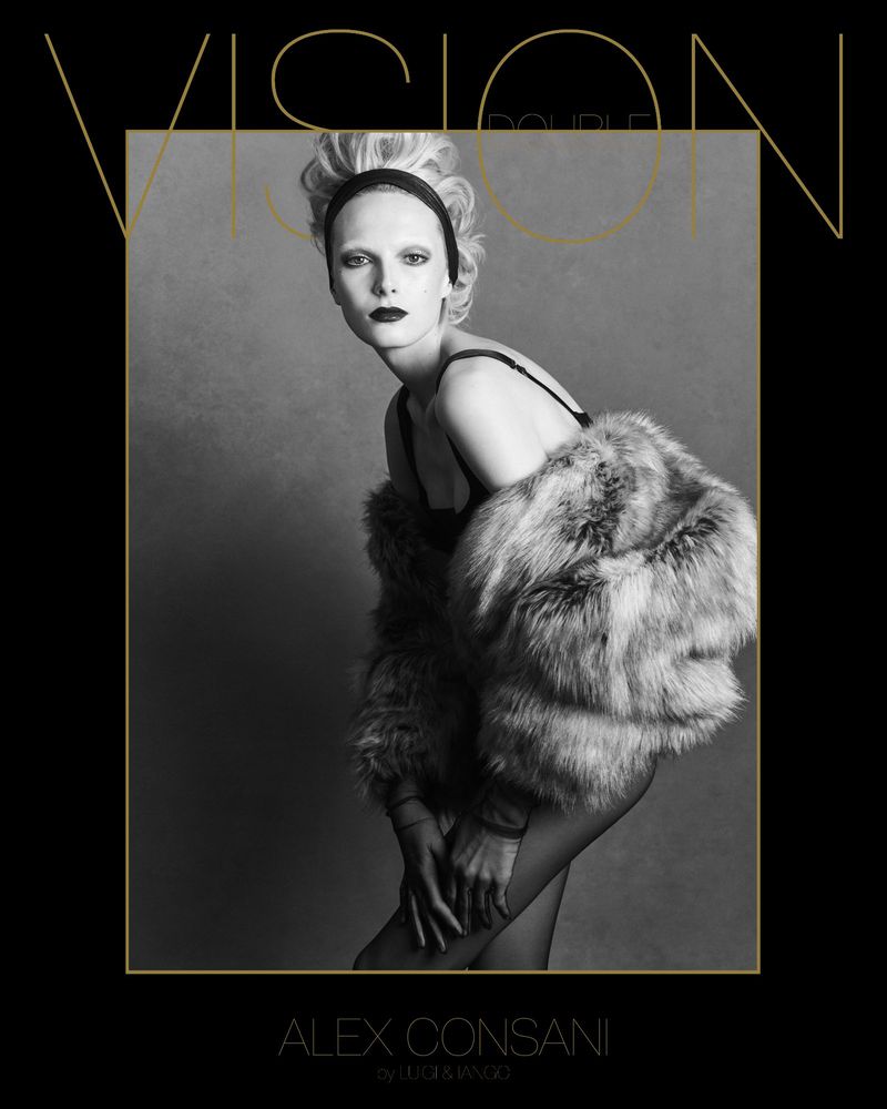 Fashion shot of an adult female with pale skin and dark lipstick wearing a fur coat and headscarf in a grey indoor environment. 