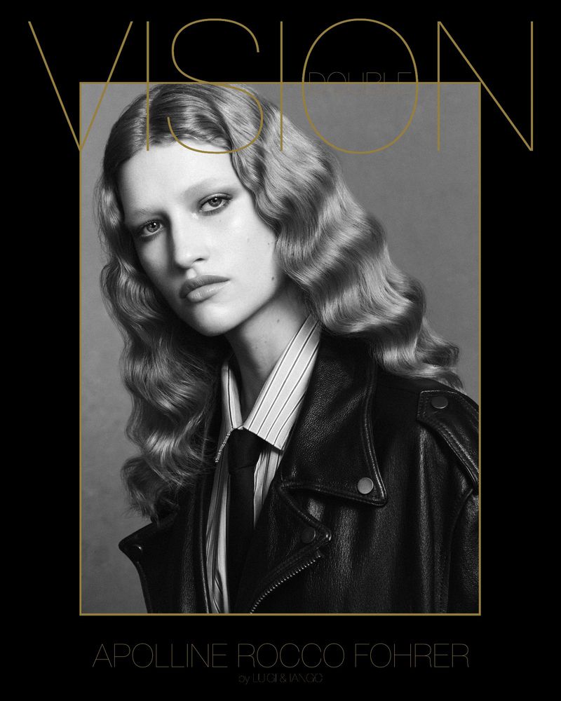 Monochrome portrait of a model with wavy hair, wearing a striped shirt, tie, and black leather jacket, against a grey backdrop.