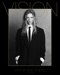 Model Caitlin Soetendal wearing a black blazer, white shirt, and black tie against a grey textured studio backdrop.