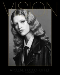Black and white portrait of a model with wavy hair wearing a black leather jacket, striped shirt, and tie against a grey backdrop