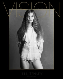 Black and white editorial portrait of a model with crimped hair wearing a lacy top against a grey textured studio backdrop.