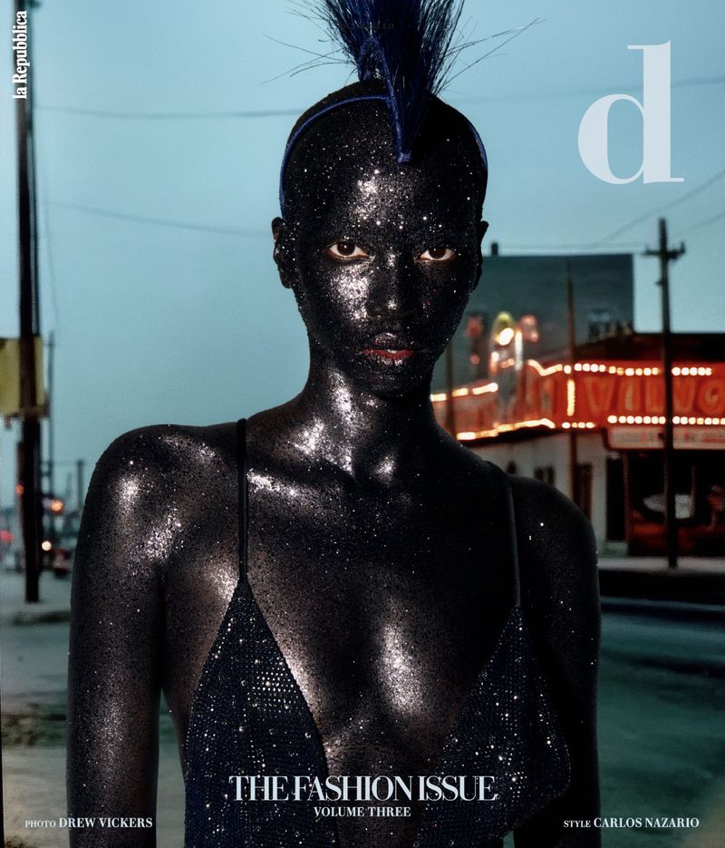 Fashion magazine cover featuring a model with black glittering skin, posing in front of a city street at dusk.