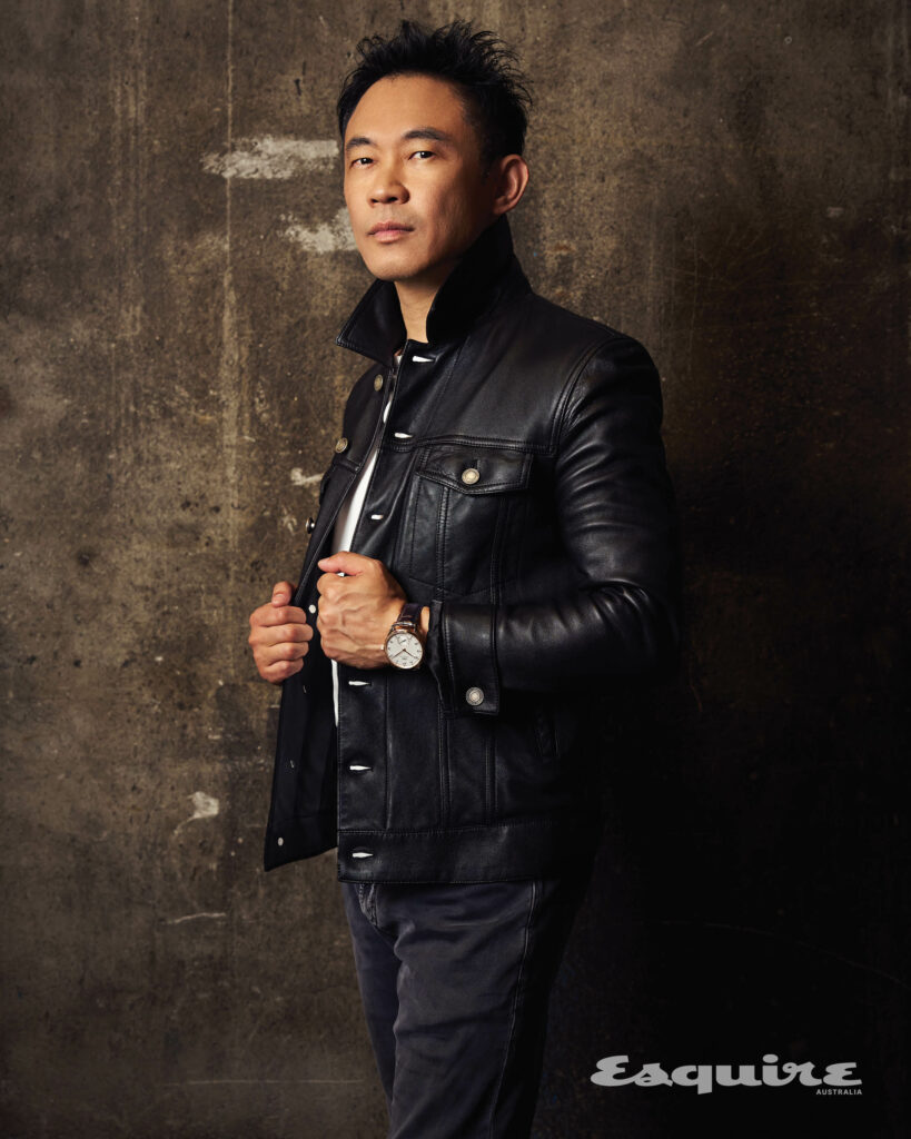 Portrait of an adult Asian man in a black leather jacket and dark jeans standing indoors against a textured wall.