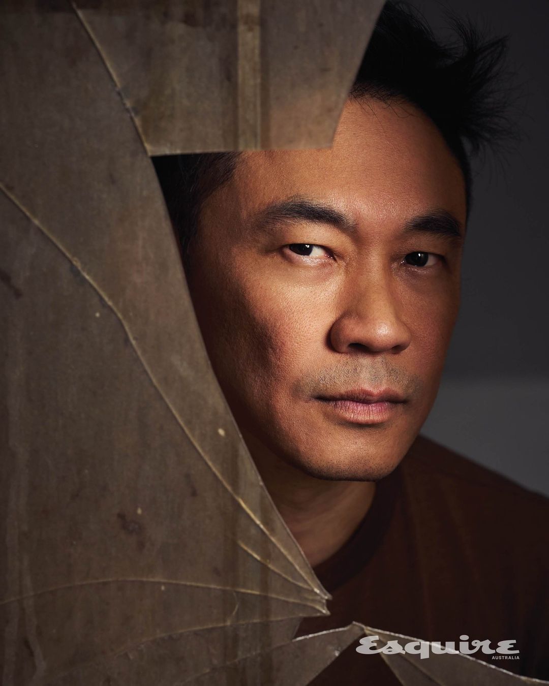 Dramatic portrait of an Asian man with short dark hair, posing indoors. He is partially obscured by shards of broken glass.