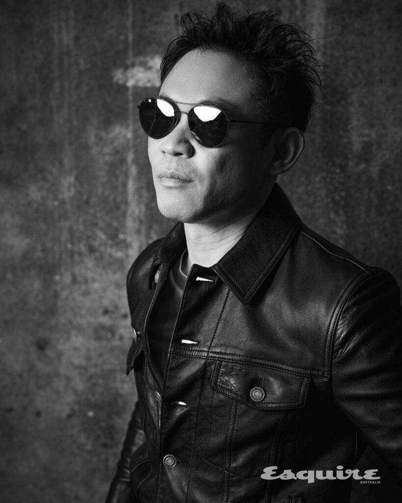 Black and white portrait of an Asian man wearing sunglasses and a leather jacket, posing in front of a textured concrete wall.