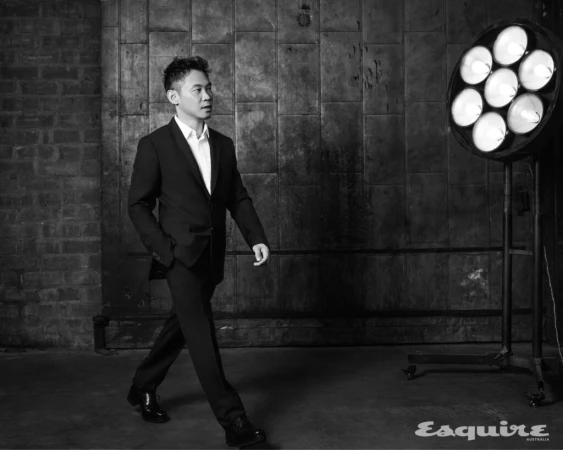 Black and white portrait of a stylish Asian man in a dark suit walking past a brick wall and studio lighting setup. Editorial style.