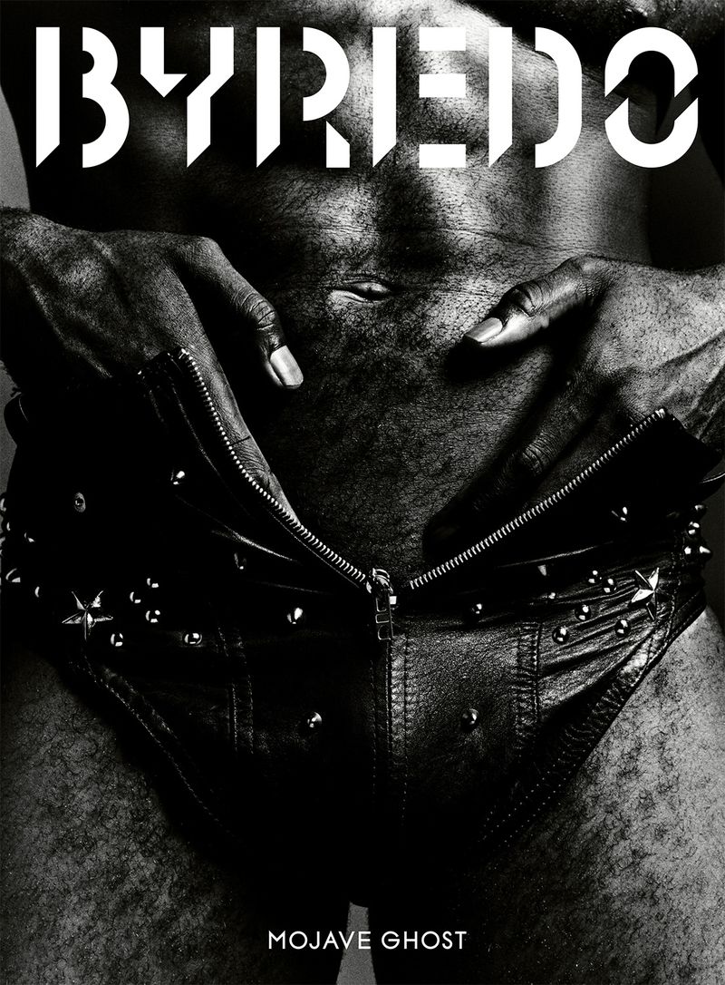 Close-up black and white shot features dark-skinned torso with hands adjusting leather studded shorts, highlighting texture and form.