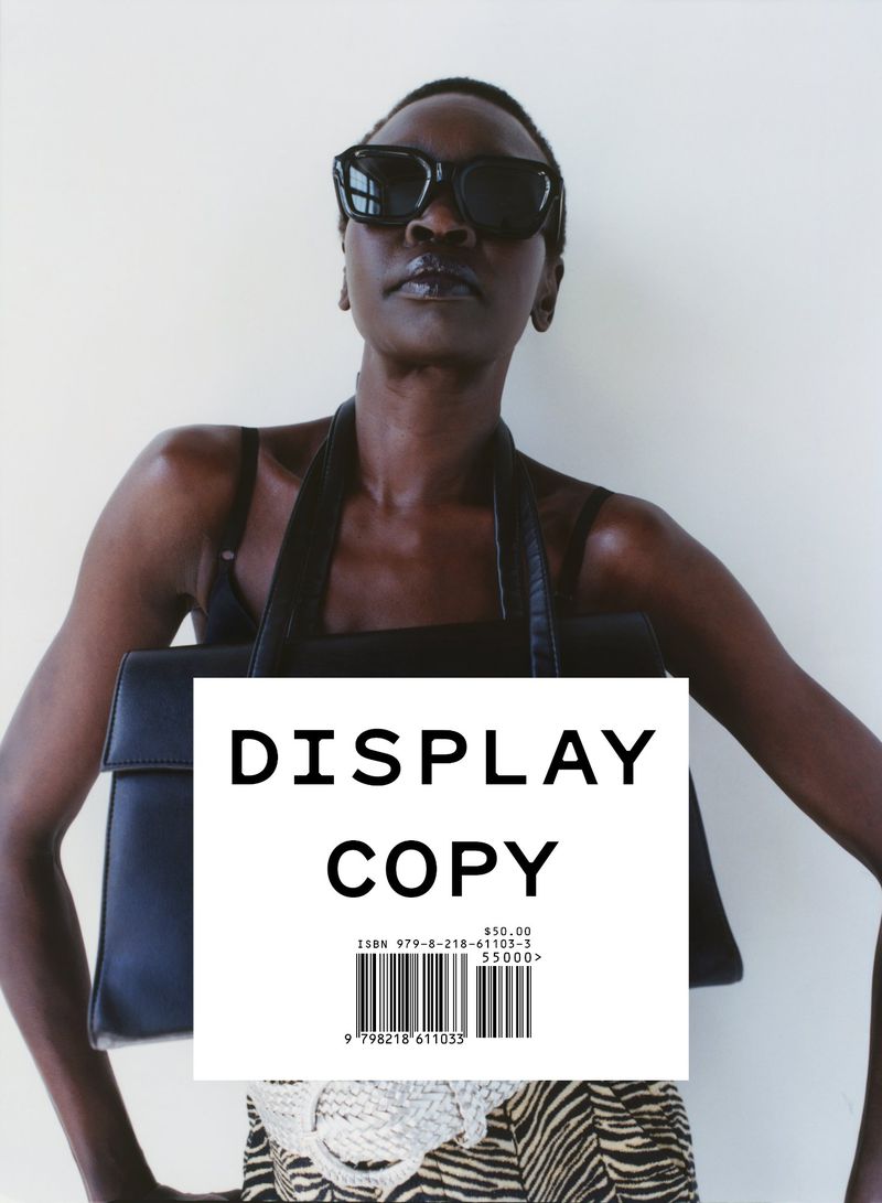Stylish woman with short hair and sunglasses poses with a black purse in a studio against a neutral backdrop, wearing a patterned skirt.