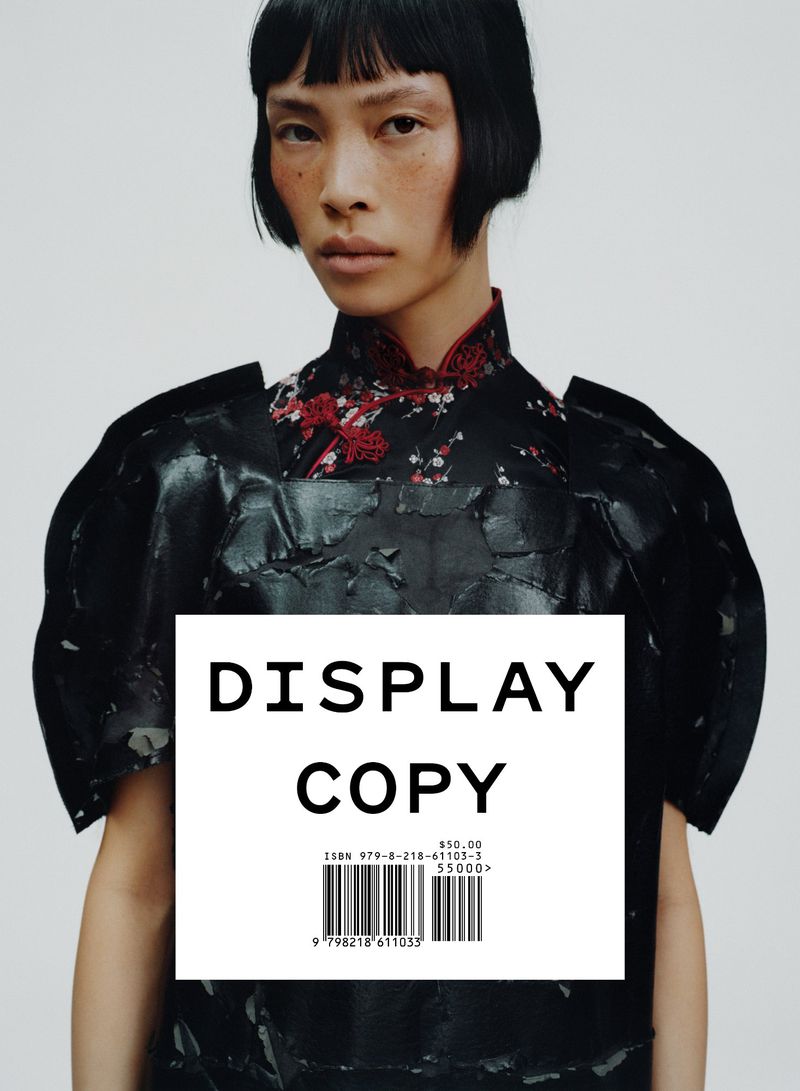 Asian model in an editorial fashion shot wearing a black textured dress with floral accents, against a pale neutral backdrop.