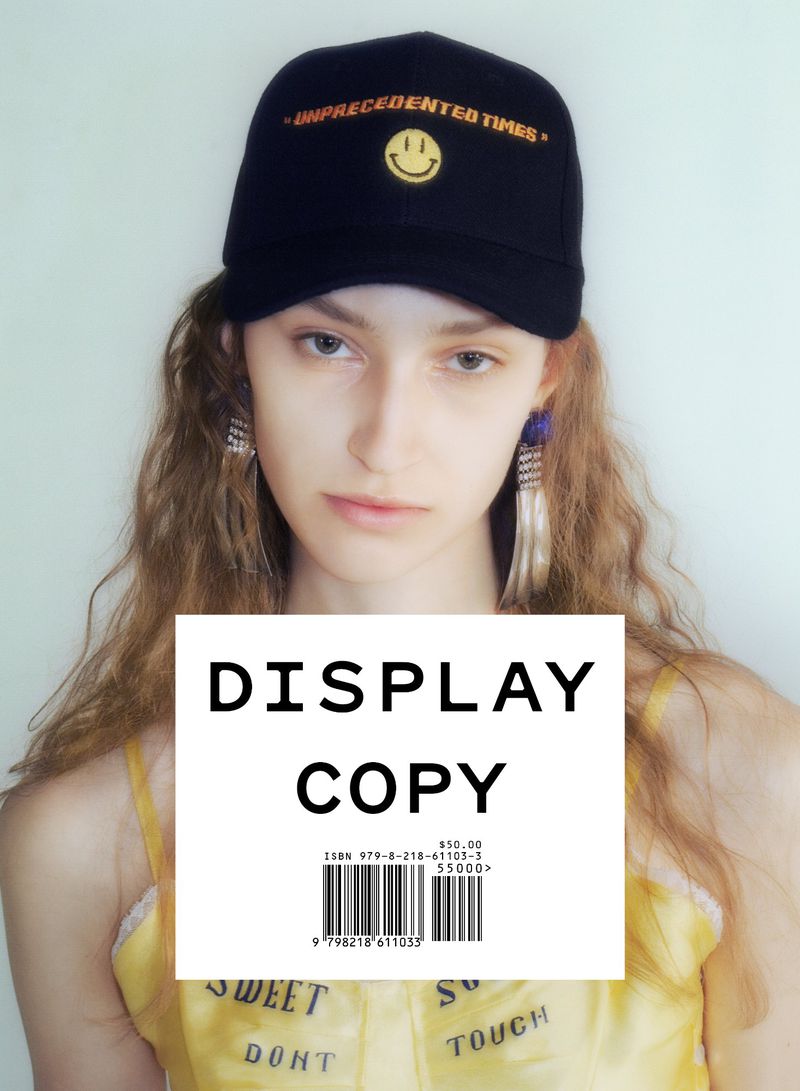 Young woman in a black baseball cap with a smiley face and the words 'UNPRECEDENTED TIMES'. She wears dangling earrings and a yellow top.