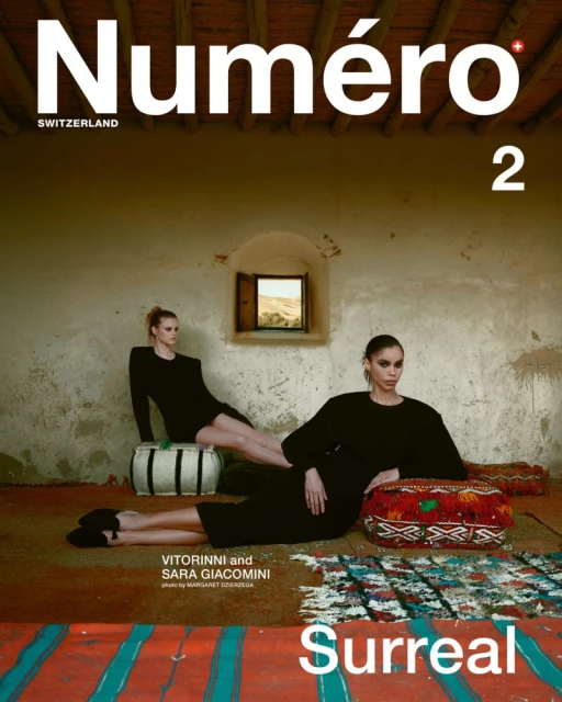 Two women in dark dresses pose indoors on a carpet with a window and textured wall for a fashion magazine cover shoot.