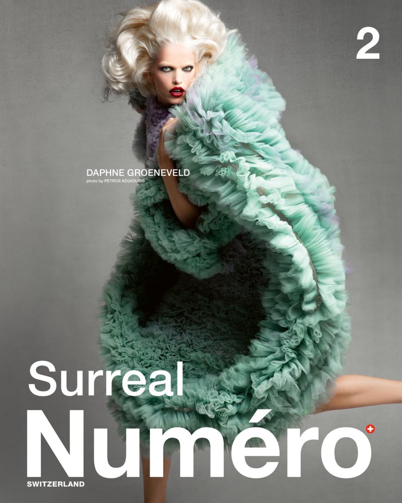 Fashion editorial cover image of a female model in a voluminous ruffled gown with fair hair posing in a studio against a grey background.