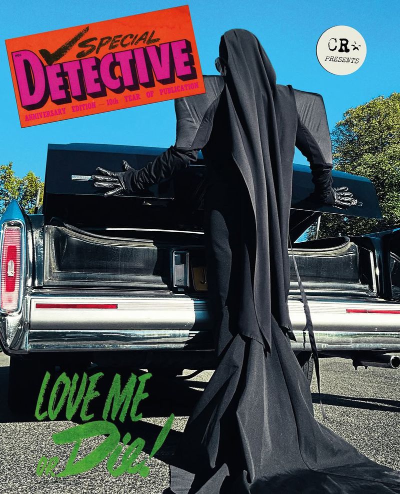 Person in black hooded robe stands at an open hearse with a magazine cover, in an outdoor setting with trees and blue sky.