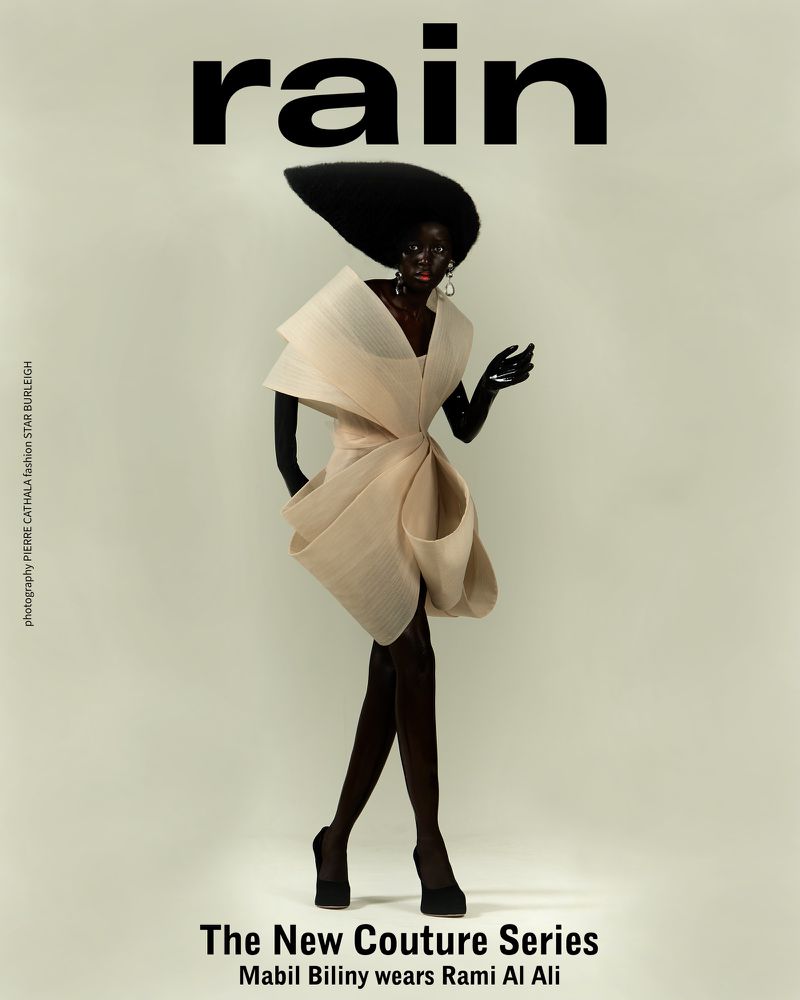 Black model Mabil Biliny poses in a Rami Al Ali couture dress with a large black hat against a neutral background for a fashion magazine.