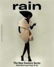 Model wearing a structured cream dress and sculptural black hat against a solid light grey background