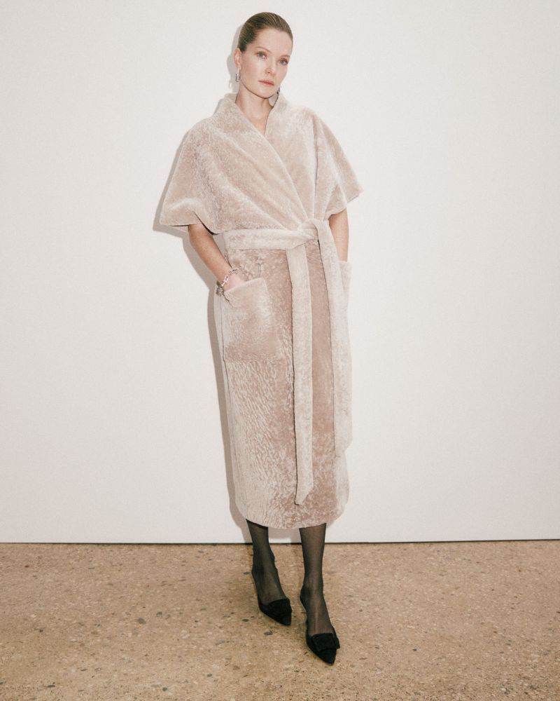 A woman in a light tan, wrap-style coat with black heels poses against a neutral wall in a studio shot, fashion editorial.