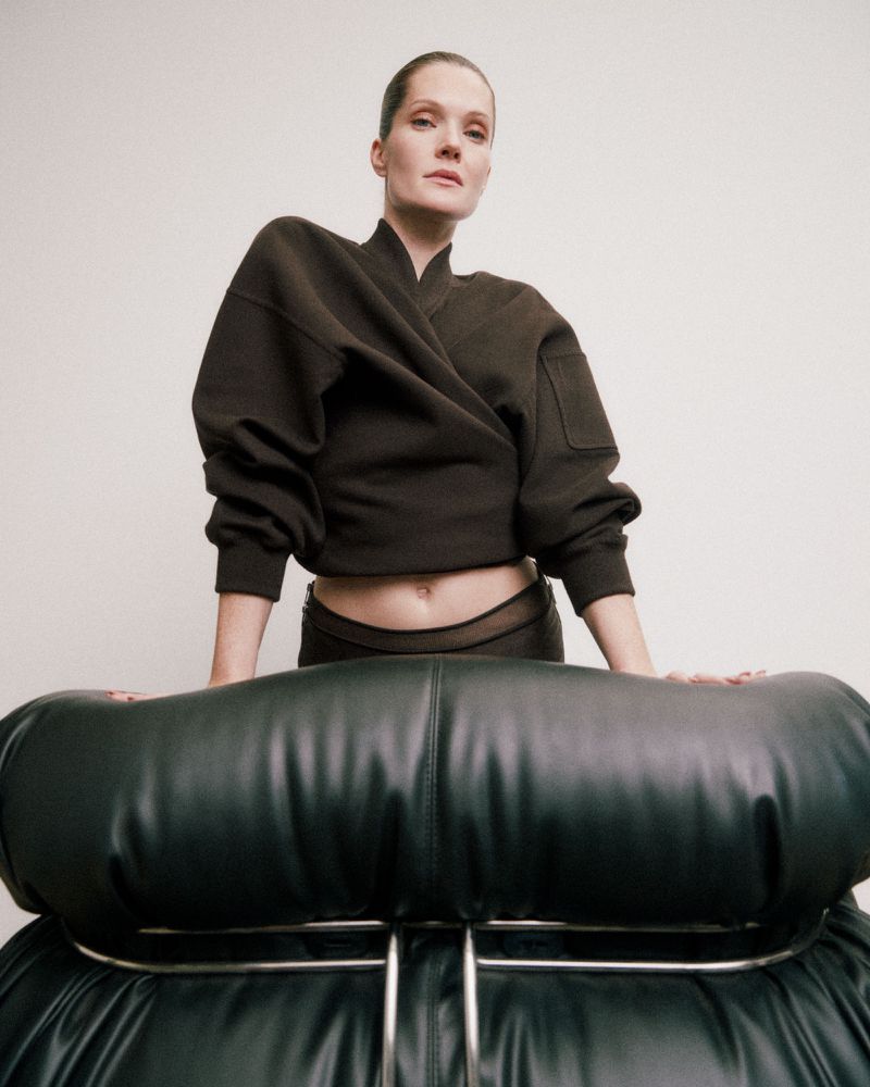 Woman in a dark-brown wrap-style top posing with a black leather chair against a white background in a studio fashion shot.