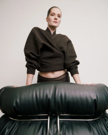 Woman in a dark-brown wrap-style top posing with a black leather chair against a white background in a studio fashion shot.