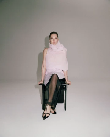 Meghann Fahy seated on a black chair wearing a pale pink draped top and sheer black skirt against a grey background