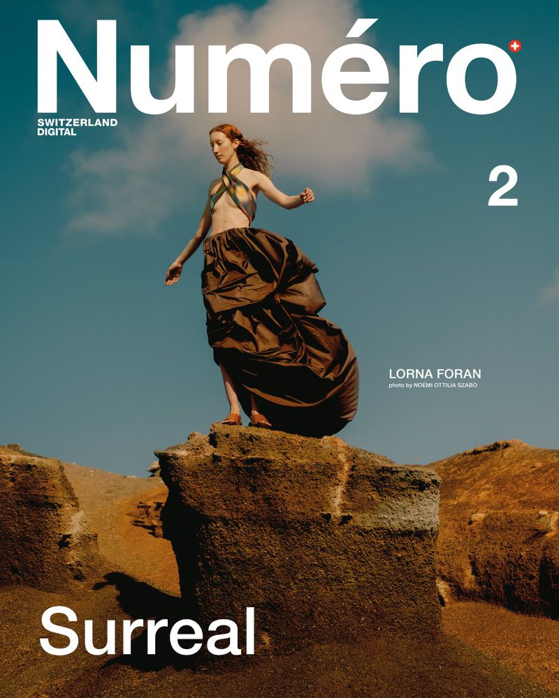 Fashion magazine cover featuring a woman in couture standing atop a rock formation against a sky with light clouds. The magazine title is prominent.