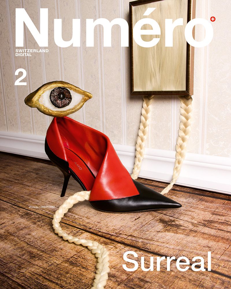Surreal magazine cover featuring a red and black high-heel shoe with an eye perched on top, juxtaposed with a framed blonde wig on a wall.