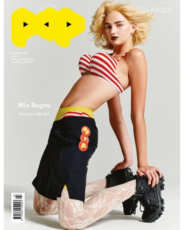 Model Mia Regan kneeling against a white backdrop wearing a striped bra top and black skirt for Pop Magazine.