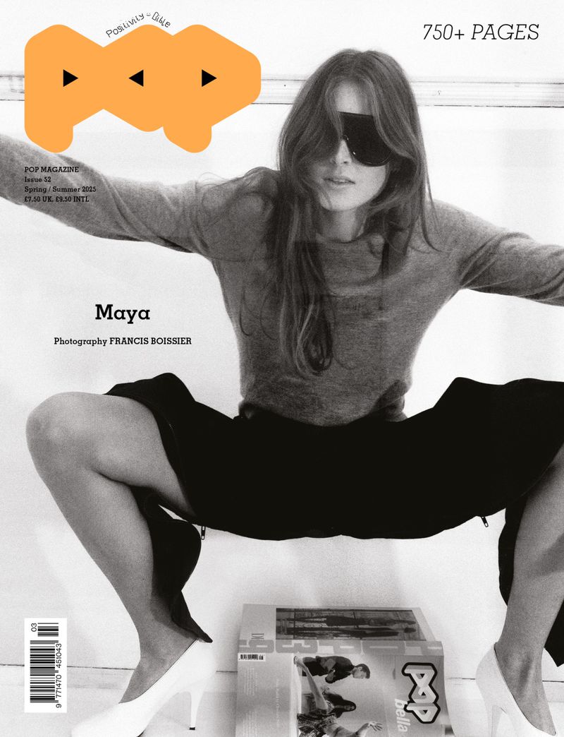 Model in grey sweater and dark sunglasses crouching over magazines with white heels against a light background