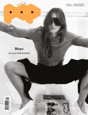 Model in grey sweater and dark sunglasses crouching over magazines with white heels against a light background