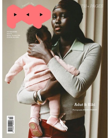 Magazine cover featuring a dark-skinned woman holding a child dressed in pink. The woman wears a grey sweater, standing against a neutral backdrop.