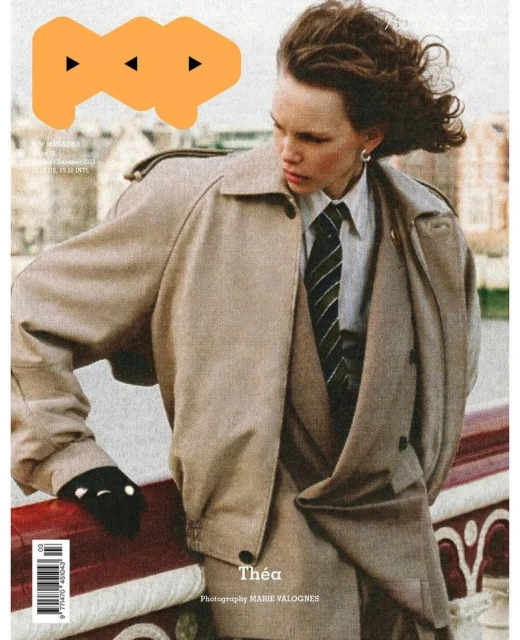 Fashion magazine cover featuring a model in a tan trench coat and suit with an orange graphic and cityscape background.