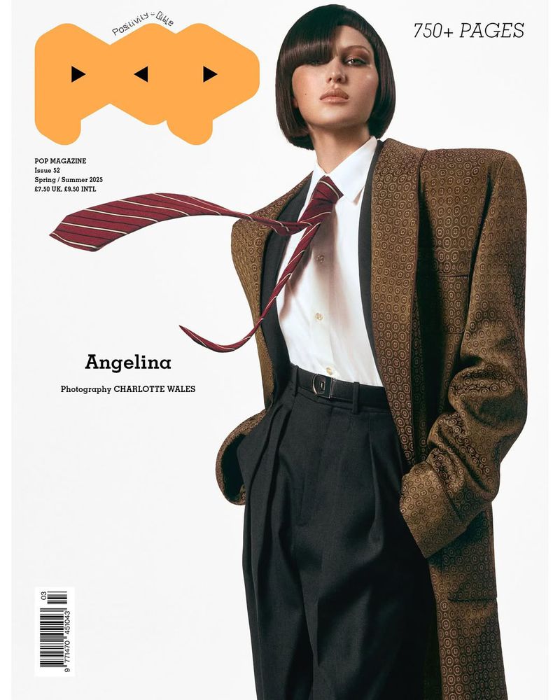 Model with dark bob hairstyle wearing structured brown blazer and blowing red striped tie against a white background.