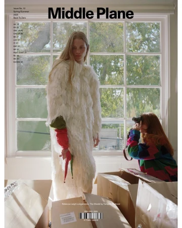 Model in white textured coat standing near large window as another person photographs her among cardboard boxes