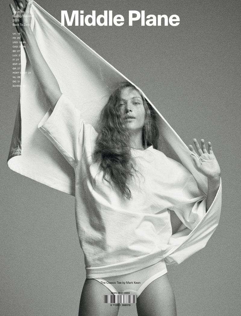 Black and white studio shot of a young woman wearing a white t-shirt and underwear, posing with a white fabric backdrop.