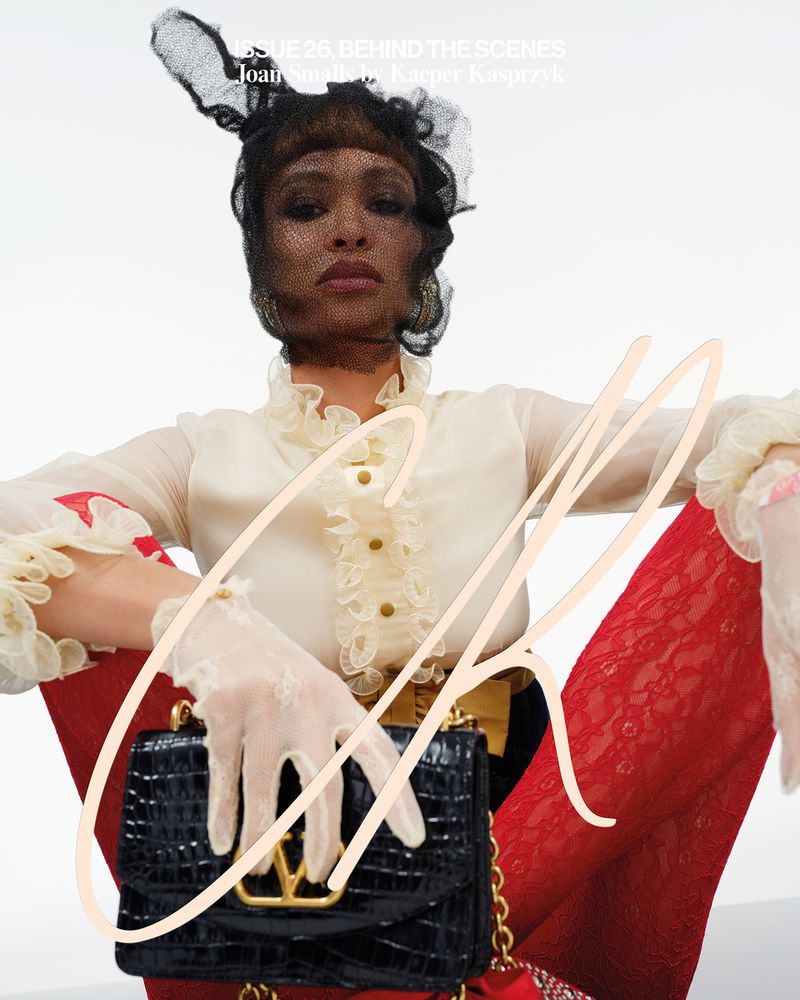 A fashion photography portrait of an adult female model wearing designer clothing and accessories against a bright, neutral background.