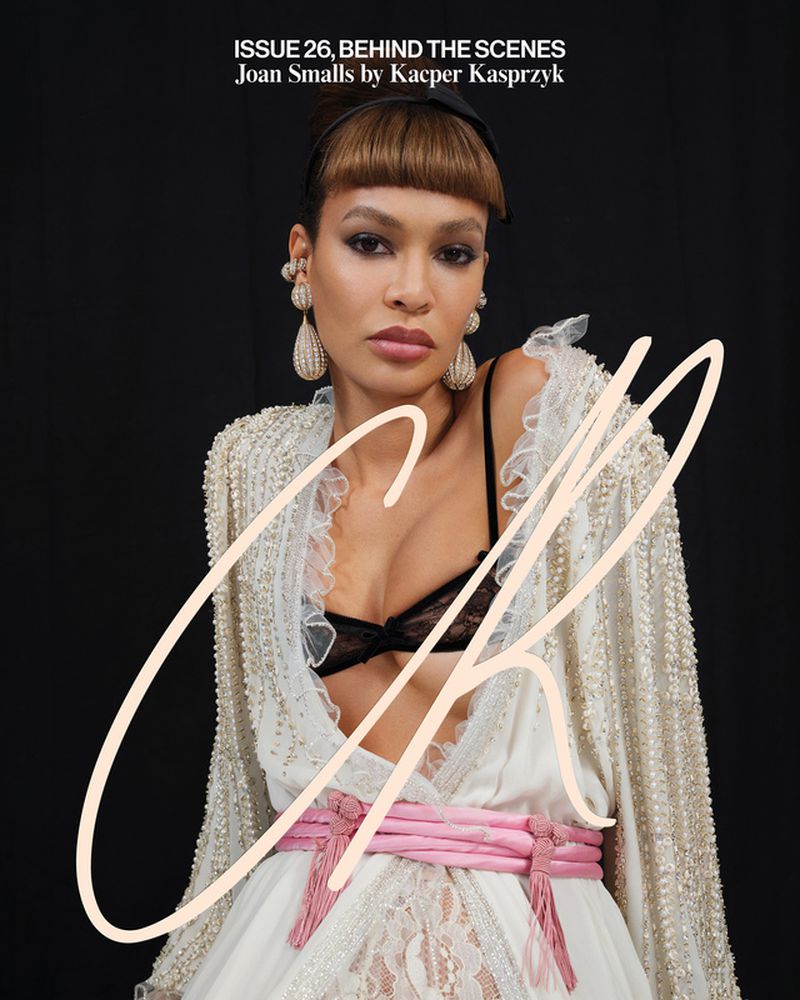 Elegant woman in an ornate white dress with a pink sash and large beaded earrings, posing against a dark background, high fashion look.