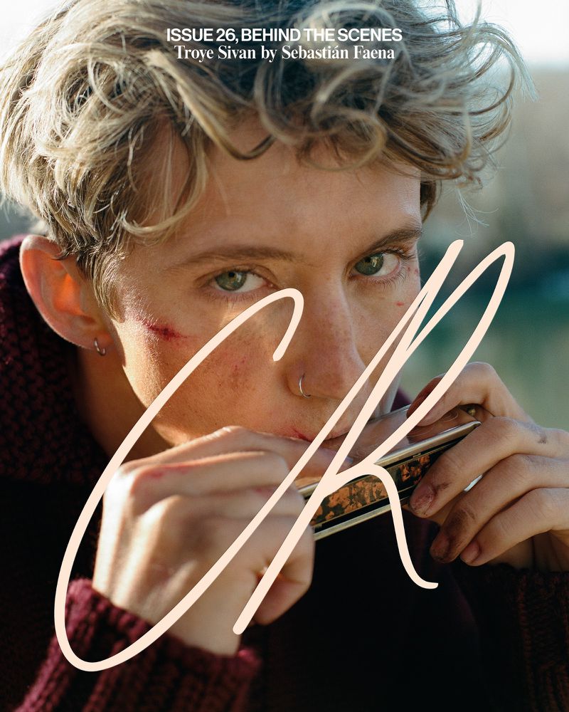 Close-up portrait of an adult male with blond hair playing the harmonica. He is wearing a maroon sweater against an outdoor background.