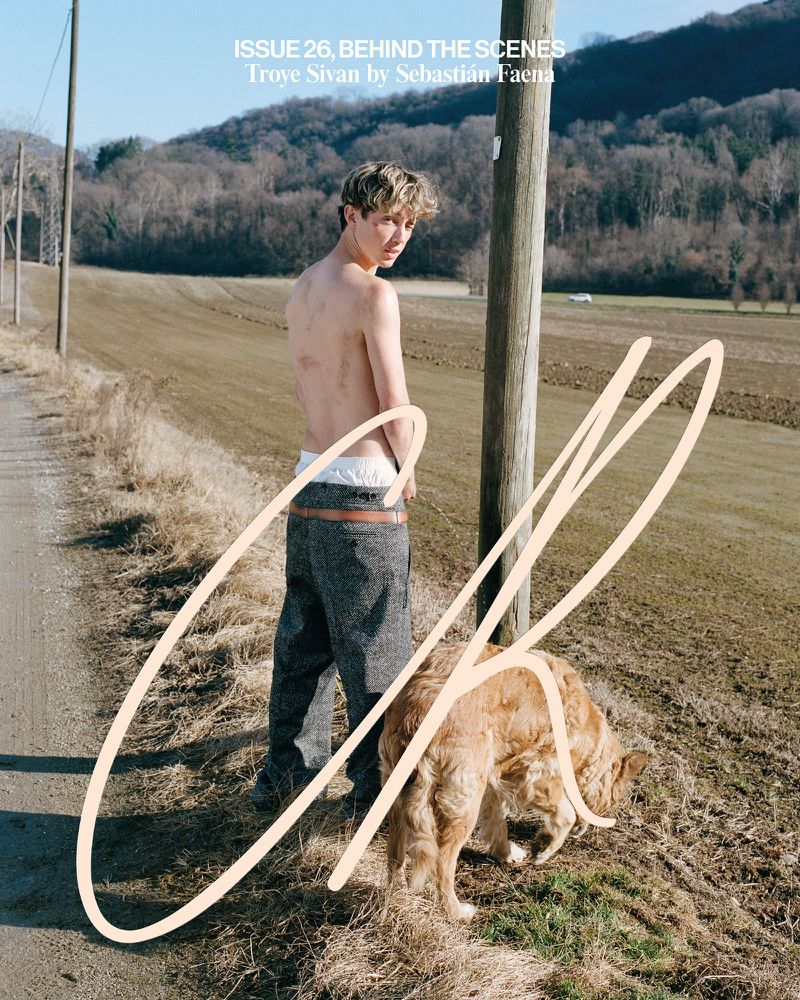 Troye Sivan, posing outdoors in casual clothing with a golden retriever dog, with a dirt road and a field in the background.