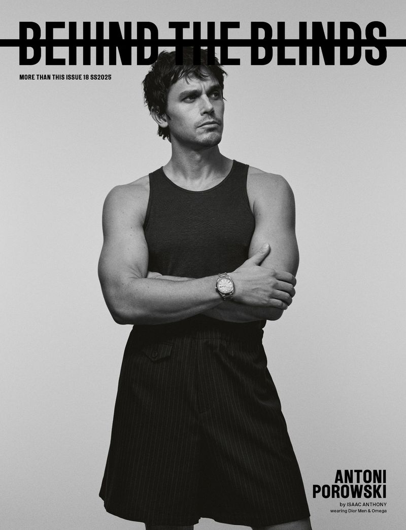 Black and white portrait of Antoni Porowski in tank top and striped skirt, arms crossed, shot for 'Behind the Blinds' magazine.