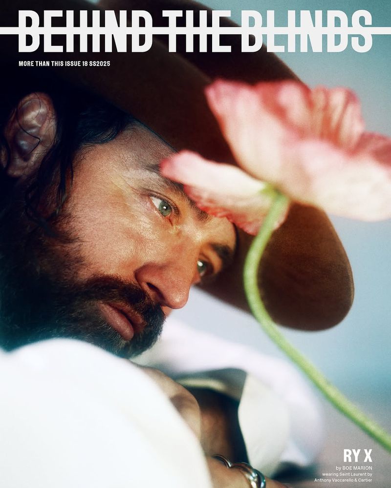 Magazine cover of a man with a beard wearing a hat and jewelry, posing with a pink flower in front of a blue background.
