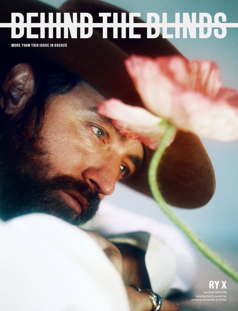 Close-up portrait of a man wearing a brown hat, looking past a large pink poppy flower held in the foreground.