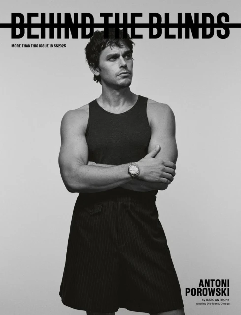 Antoni Porowski wearing a dark tank top and dark shorts with arms crossed against a plain grey background