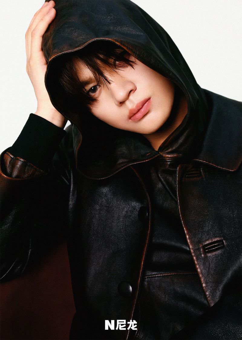 Young adult with hand on head wears a dark hooded leather jacket in a portrait with neutral background. Fashion and beauty shot.