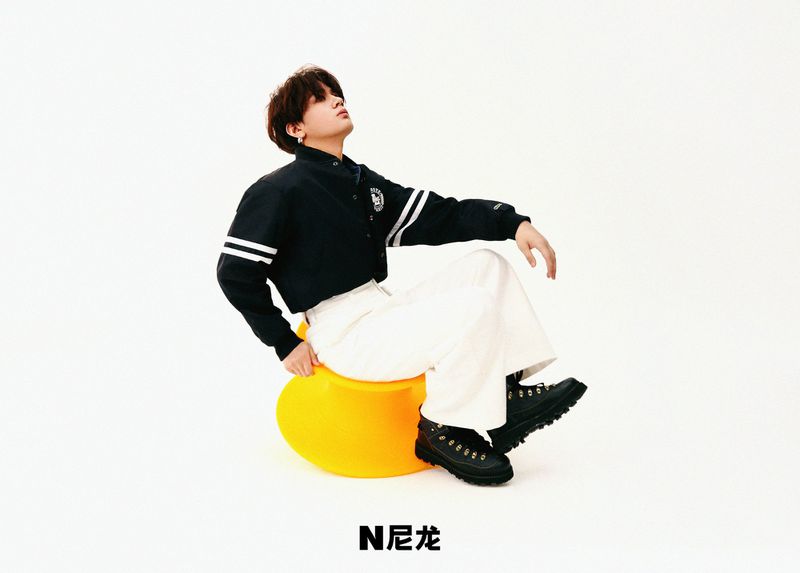 Young adult poses on a yellow ball seat in a photo studio wearing a black and white jacket, white pants, and black boots.