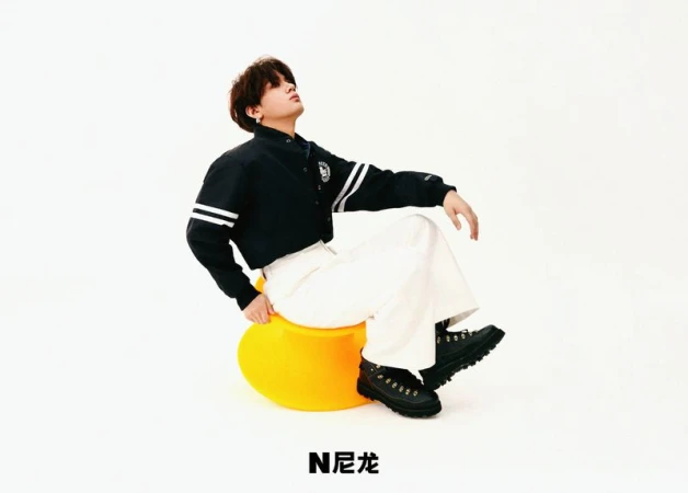 Young adult poses on a yellow ball seat in a photo studio wearing a black and white jacket, white pants, and black boots.