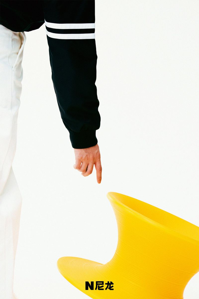 Person's hand points towards a yellow modern chair. The person wears a black shirt with white stripes and white pants in a white space.