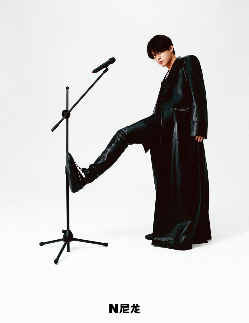 Person in black leather coat and boots stepping on microphone stand in studio. Stylish studio portrait on white backdrop.