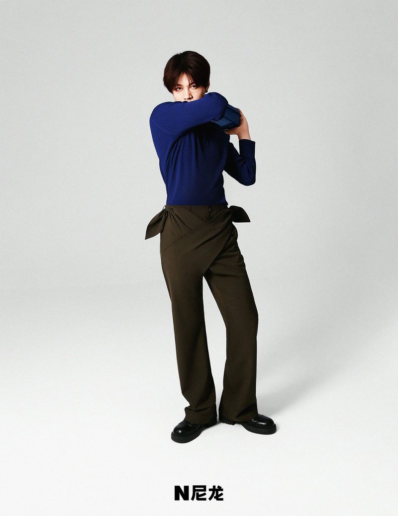 Fashion shot of a model in a blue long-sleeved shirt and olive green pants, standing in a light grey studio with brand logo.