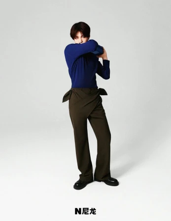 Fashion shot of a model in a blue long-sleeved shirt and olive green pants, standing in a light grey studio with brand logo.