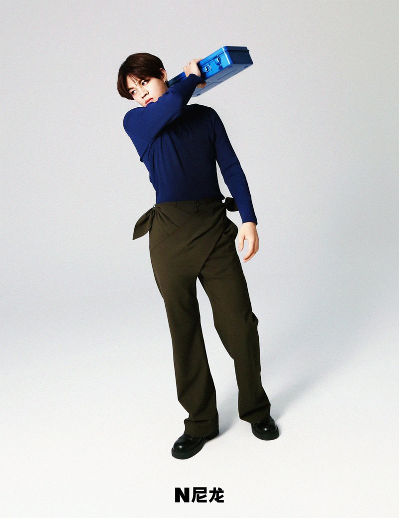 Adult man in stylish blue shirt and olive green wrap pants poses while holding a blue suitcase in a studio setting against a white backdrop.